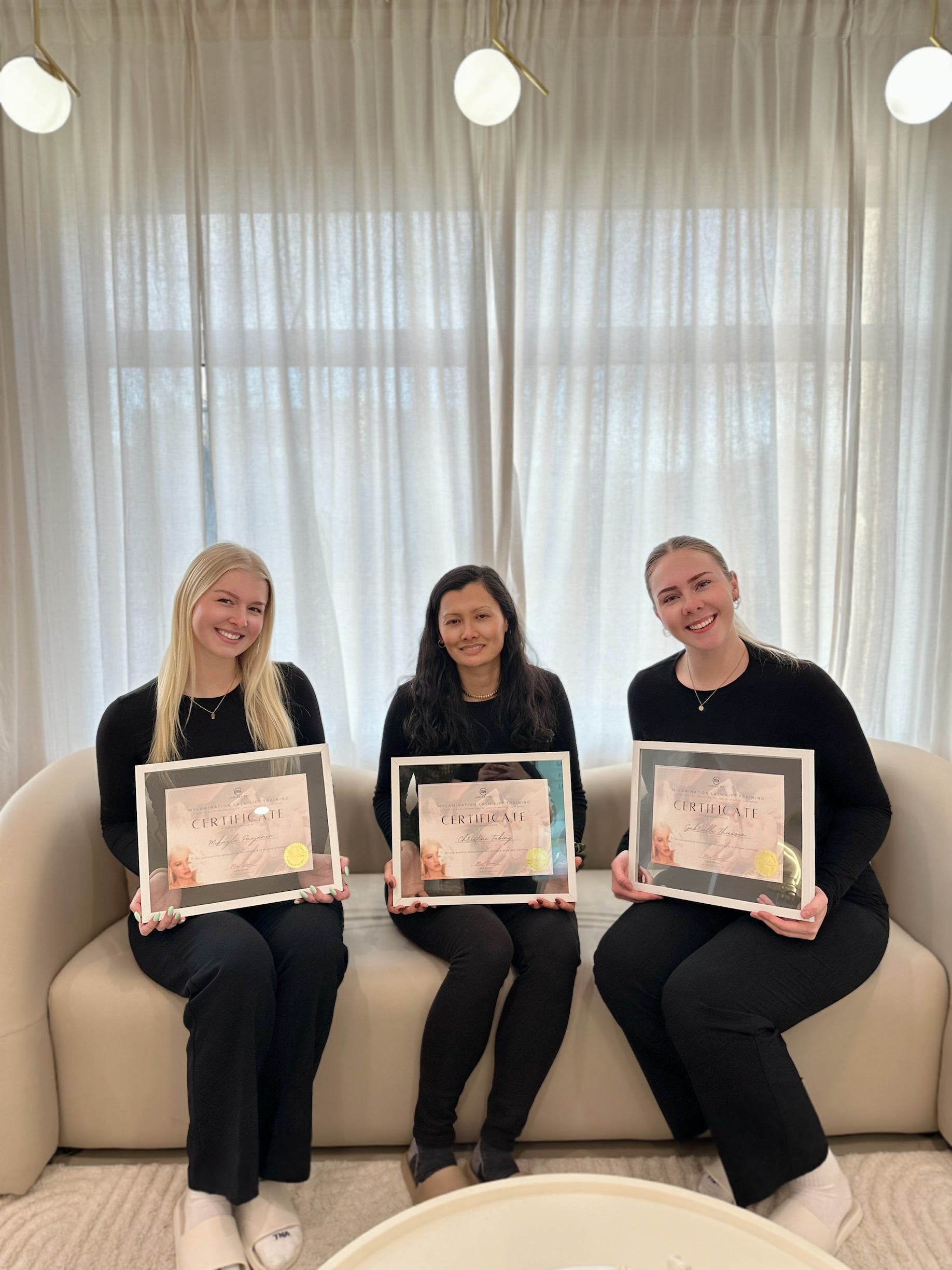 Three women sitting on a couch holding framed certificates in a modern interior setting.